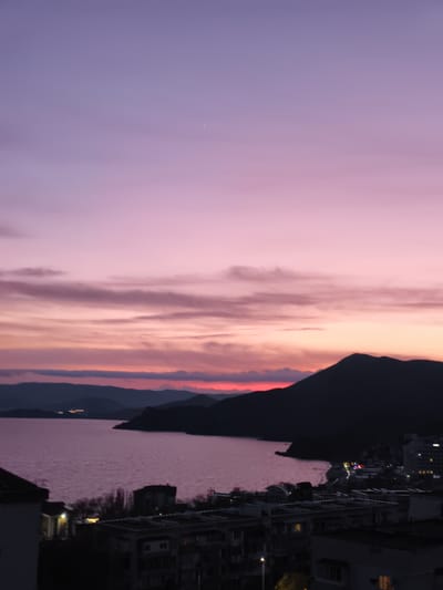 A scenic view captures a city situated alongside a body of water, likely the sea, under a twilight sky. In the foreground, the city's architecture is visible, comprising apartment blocks and buildings with illuminated windows, indicating the presence of residents. The waterfront is softly illuminated, with scattered lights reflecting on the water's surface.

In the middle ground, the sea extends, bordered by a landmass or peninsula that juts out, silhouetted against the fading light of the sunset. The sky transitions from a deep purple at the zenith to a warmer, reddish hue near the horizon, punctuated by scattered clouds that catch the last rays of the sun.

The image suggests a tranquil evening in the city, with the natural beauty of the sunset contrasting against the urban environment. The location is speculated to be within the vicinity of the Орджоникидзевский поселковый совет region of Ukraine, based on known landmarks and characteristics.