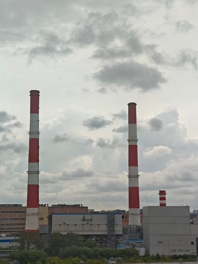 This media file shows an industrial scene with multiple smokestacks against a cloudy sky. The main subjects are three tall, cylindrical smokestacks. Two of the smokestacks are prominently featured in the foreground, painted with alternating red and white bands. The third, smaller smokestack is located in the background, also with red and white banding.
Behind the smokestacks, a complex of industrial buildings is visible. These structures are varied in color and design, with some featuring large, rectangular windows and others appearing more modern with metallic siding. Green foliage, likely trees and bushes, forms a dense layer in the lower part of the frame, suggesting the industrial area is bordered by natural elements or a park.
The sky is overcast with a significant amount of grey clouds, indicating a possibly cool or overcast day. The lighting suggests it is daytime, but the cloud cover diffuses the light. There are no visible people or specific activities taking place. The overall mood is industrial and somewhat somber due to the weather and the stark industrial structures.
Location cues, such as the style of architecture and the presence of large smokestacks, align with an industrial zone in Moscow, Russia. No text is discernible in the image. The scene depicts a power plant or a similar industrial facility.