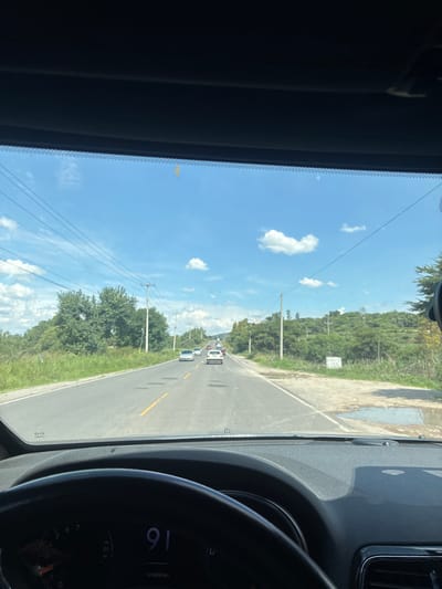 A first-person perspective view from inside a vehicle shows a two-lane road under a bright blue sky with scattered clouds. The road is asphalt and has yellow lines marking the lanes. Several vehicles are visible on the road ahead, and trees and vegetation line the sides. The dashboard of the vehicle is partially visible, including a speedometer displaying "91". The location is identified as Santiago Tlautla, Mexico.