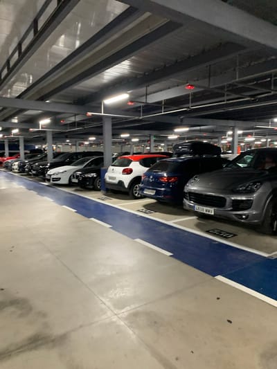 The image depicts a dimly lit multi-story parking garage, likely located in Manises, Spain, as indicated by the license plate "4838 MMV" which is consistent with Spanish plates. The perspective is from the driver's seat of a parked car, looking forward and to the left. Rows of cars are parked in designated spaces, separated by blue painted lanes and white lines. The ceiling of the garage is visible, featuring metal beams and fluorescent lighting. Some red emergency lights are also discernible on the ceiling. The concrete floor shows signs of use, with some staining and tire marks. Several vehicles are clearly visible, including a dark gray Porsche Cayenne in the foreground with license plate "4838 MMV", a dark blue hatchback with a roof box, and a white hatchback with a red stripe. Further down the row, other cars are parked, mostly dark sedans and hatchbacks. No people are visible in the frame. The overall impression is one of a typical, functional, and somewhat sterile parking facility.