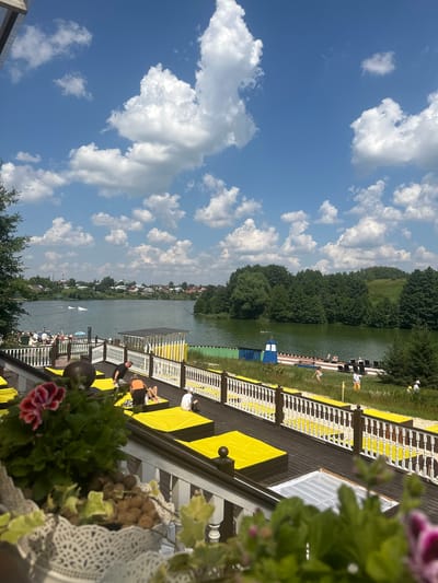 This media file depicts a sunny day scene by a body of water, likely a lake or river, at Dal'nie Prudishchi, Russia. The perspective is from a wooden deck with yellow and white accents, overlooking the water.
The primary subjects are people relaxing on the deck and in the water. On the deck, several individuals are seated or lying down on yellow mats, enjoying the sunshine. Some appear to be sunbathing or socializing. In the water, a few people are visible, possibly swimming or engaged in water activities. There are also small boats on the water.
The scene is set on a wooden pier or deck with a railing. Yellow mats are spread out on the deck, creating a vibrant visual element. Green grass borders the deck and slopes down towards the water. A line of trees forms the background on the opposite bank, with some houses visible in the distance, suggesting a semi-rural or recreational area.
The weather appears to be clear and warm, indicated by the bright sunshine and scattered white clouds against a deep blue sky. The time of day is likely midday or early afternoon, given the strong sunlight. There is no visible text in the image. The overall mood is one of leisure and relaxation on a summer day.