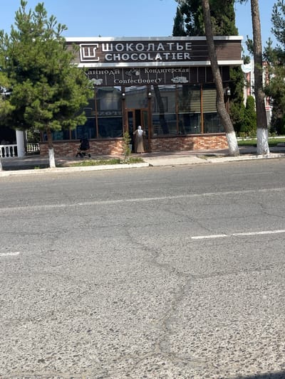 The image shows a street scene in the city of Ismoili Somoni, Tajikistan, featuring a confectionary shop. The shop's facade is adorned with text in both Cyrillic and Latin scripts, reading "ШОКОЛАТЬЕ" and "CHOCOLATIER". Below this, further text appears, with "Кондитерская" and "Confectionery" visible. A person wearing a headscarf and a long dress is entering the shop. To the left of the shop, a black stroller is parked on the sidewalk. The street in front of the shop is paved with asphalt, and trees line the sidewalk. The sky is clear and blue.