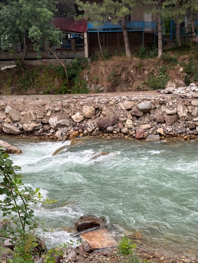 The image depicts a fast-flowing, turquoise river in Khumsan, Uzbekistan. The riverbanks are lined with a mixture of rocks and gravel. Above the river, on the embankment, is a structure that appears to be a building with a red metal roof and blue railings. Trees and vegetation are present on the embankment and surrounding the building. The scene is naturalistic, with the river being the dominant feature. There are no people or animals visible in the image. The lighting suggests it is daytime.