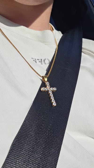The image shows a close-up of a person wearing a white t-shirt and a gold chain with a cross pendant adorned with clear stones. The pendant is resting on a dark blue seatbelt. The t-shirt has some faint, dark text on it that appears to be "OFF". The person's arm and shoulder are visible. The lighting suggests it is daytime, with sunlight casting shadows. The scene is likely inside a vehicle, given the seatbelt. The setting is not explicitly identifiable beyond this context. There are no overt emotional cues visible, but the focus on the jewelry and the casual attire suggest a personal or everyday moment. The location context of Liliak, Bulgaria, does not provide specific visual cues within the image itself.