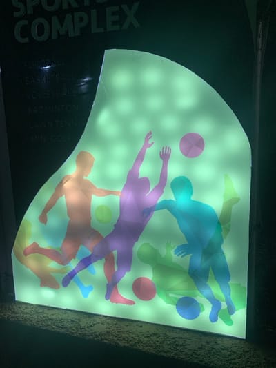 A brightly lit sign for a sports complex features various athletes in translucent hues of blue, purple, orange, green, and yellow, playing with balls. The silhouette of players creates an overlapping effect. A dark border of the sign lists the sports offered: football, basketball, volleyball, badminton, lawn tennis, and mini golf. The illuminated background appears to be made up of numerous individual lights, shining through the image.