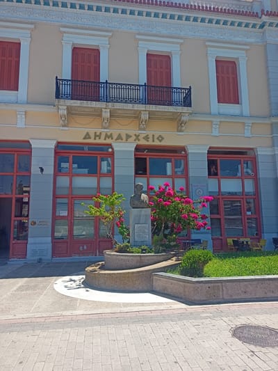The image shows the front of the Town Hall in Chalkida, Greece. The building is painted a pale yellow with ornate white detailing around the windows and roofline. The windows on the upper floor have red shutters. A black wrought-iron balcony with a decorative railing spans the facade above the ground floor. The ground floor features large, red-framed windows and doors. A statue of a bust on a pedestal stands in front of the building, to the left of center, partially obscured by a flowering bush with bright pink blossoms. A plaque is visible on the ground to the left of the statue, and another, larger plaque is mounted on the building's facade to the left of the entrance. The area in front of the building is paved with light-colored stone tiles and features concrete planters with greenery. Tables and chairs are visible through the ground floor windows, suggesting a cafe or seating area inside. The sky is clear and blue, indicating a sunny day.