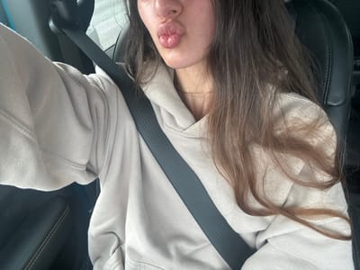 A young woman with long, brown hair and light skin is sitting in the driver's seat of a car, wearing a light-colored hooded sweatshirt and a thin gold necklace. Her lips are pursed in a kiss. A black seatbelt is fastened across her chest. The interior of the car appears to be black leather. The background is blurry, but suggests an outdoor environment. The image was taken from the woman's perspective.