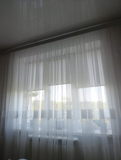 The image shows a view through sheer white curtains, revealing a windowsill with several potted plants. The plants are of varying sizes and types, some appearing to be succulents or small leafy plants. Beyond the windowsill, a bright, sunlit scene of greenery and trees is visible, suggesting it is daytime. The light filtering through the curtains creates a soft, diffused illumination, hinting at pleasant weather. The curtains themselves are a prominent feature, with a textured band visible near the bottom. In the lower portion of the image, the top of a radiator can be seen, indicating the scene is likely indoors, in a room with a window. The overall impression is one of a peaceful, domestic interior looking out onto a bright, natural landscape. There is no visible text in the image.