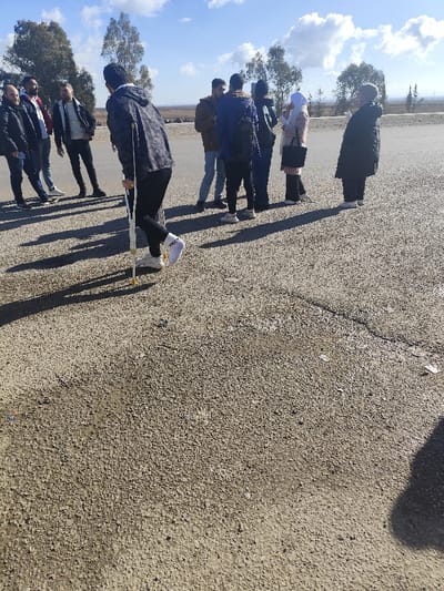 A group of people stand on a paved surface under a blue sky with scattered clouds. One person walks using crutches. Shadows are cast by the individuals and objects, suggesting bright sunlight. Trees are visible in the background. The foreground consists of the textured pavement. The group appears to be casually gathered, possibly observing something off-camera.