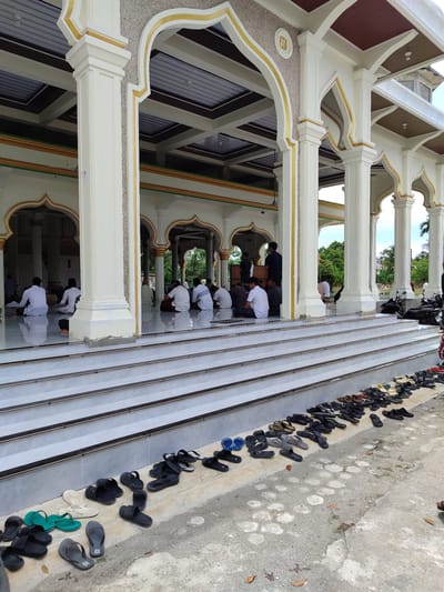 The image depicts the exterior of a mosque in Lancoklancok, Indonesia. The mosque features prominent white pillars with gold accents, archways, and a tiered white staircase leading up to the entrance. The scene is filled with numerous pairs of sandals and flip-flops arranged neatly on the ground and on the steps, indicating that people have removed their footwear before entering the mosque.

Several people are visible inside the mosque's covered area, seated on the floor in prayer or quiet contemplation. They are dressed in modest attire, predominantly white clothing and head coverings. The overall atmosphere appears to be one of peaceful observance.

The time of day is likely daytime, given the bright illumination. The weather appears to be pleasant, with a clear sky visible in the background. The setting is clearly religious, given the architecture of the mosque and the presence of people engaged in prayer. The abundance of shoes suggests a gathering or a period of worship. There is no visible text in the image.
