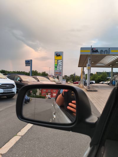 The image depicts a view from inside a vehicle, looking towards a gas station. In the foreground, a side-view mirror reflects a person's hand and arm. Beyond the mirror, the gas station, identified as an Eni station by its signage and yellow canopy, is visible. A tall price display shows fuel costs, with numbers like 1.889, 1.989, 1.979, 2.039, and 2.239. Several cars are lined up at the pumps. A Ford service sign is also visible. The scene appears to be in Pegnitz, Germany, suggested by the presence of European car models and the overall architecture and landscape. The sky is partly cloudy with a hint of sunset colors.