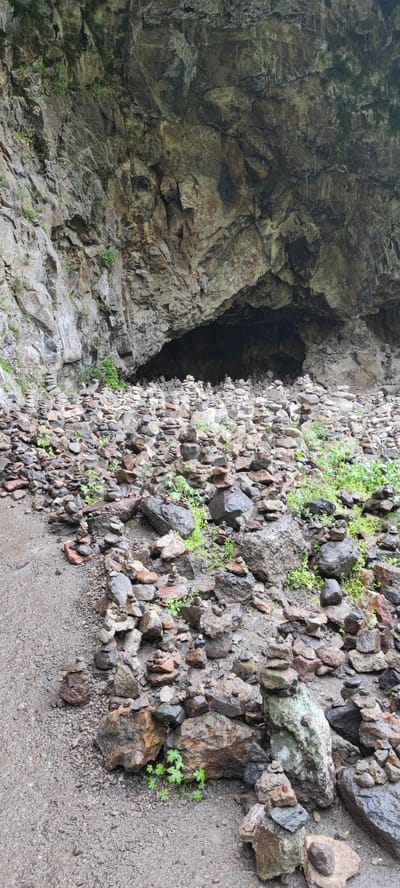 The image depicts a site in Goght, Armenia, characterized by a large rock overhang or shallow cave, in front of which are numerous small stacks of stones, each constructed of several rocks balanced on top of one another. The ground is covered in similar loose stones and some sparse green vegetation. The overall impression is of a rocky and damp environment. There are no people visible in the scene. The primary action appears to be the intentional stacking of stones.