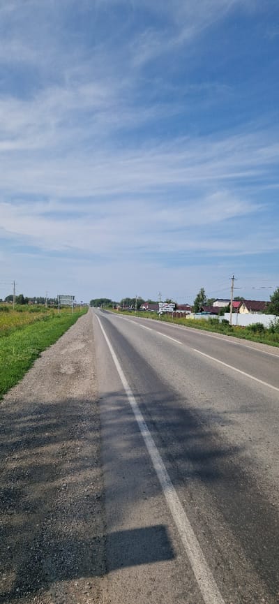 The image depicts a wide, paved asphalt road in the countryside, likely near the city of Leninskoye, Russia. The road has a white painted line marking the lanes and a gravel shoulder on the left side. To the right of the road, there are several houses with red tiled roofs and fences, suggesting a residential area or village. Beyond the houses, trees and vegetation are visible, and further in the distance, more trees line the horizon. The sky is blue with scattered white, wispy clouds. There are power lines and utility poles along the side of the road, and a few vehicles can be seen in the distance on the road. A sign with text is visible near the right side of the road, though the text is not legible in detail. The overall impression is of a sunny day in a rural or semi-rural setting.