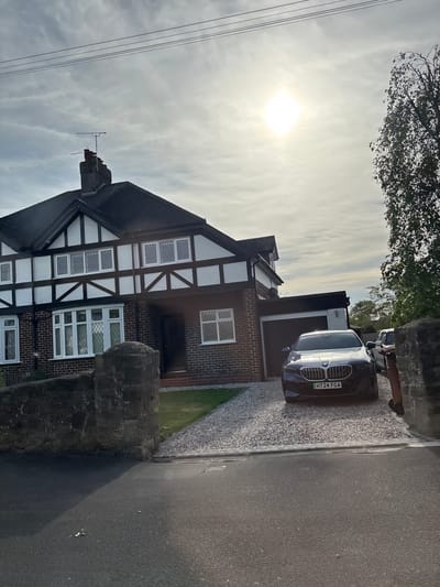 The image shows a two-story Tudor-style house in Wistaston, United Kingdom. The house has a black roof, white stucco siding with dark brown timber framing, and multi-paned windows. A dark-colored BMW with license plate "HT24 FGA" is parked in front of the house on a gravel driveway. A brick garage is attached to the house. A gray paved road is in the foreground, bordered by a low stone wall. Overhead power lines are visible on the left side of the image. The sky is overcast, with the sun partially obscured by clouds. A tree is in the upper portion of the image.