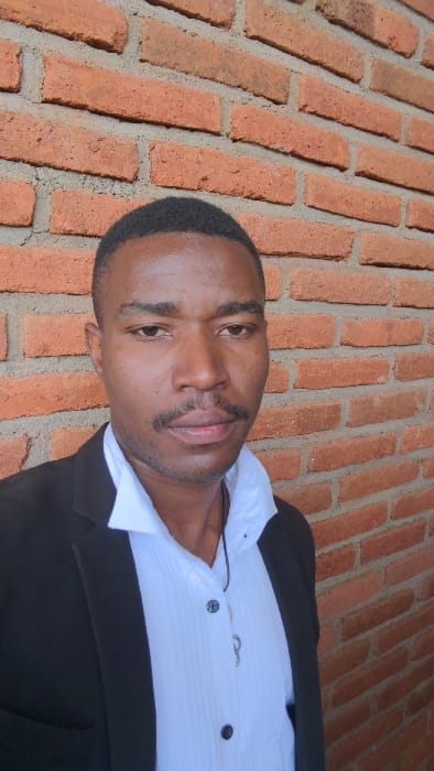 A close-up image showcases a dark-skinned man wearing a crisp white shirt and a black blazer. His facial features are clearly visible, including his mustache and a slight smile. The background is a textured brick wall, adding depth and contrast to the shot. The portrait focuses on the man's face, highlighting his features against the backdrop.