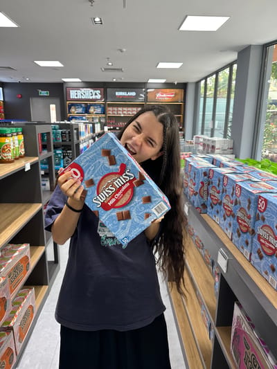 A young woman stands in a store aisle, holding a box of "Swiss Miss Milk Chocolate Flavor Hot Cocoa Mix (50-1.38 oz envelopes)." The aisle features shelves filled with products, including other boxes of Swiss Miss and Nissin Cup Noodles. The brand "Hershey's" is visible in the background, as is "Kirkland". The store has modern lighting and polished floors. The woman is grinning, and her dark hair is styled with braids near her face. The location is a store, potentially a Costco. It's not possible to confirm that the location is in Ben Cau, Vietnam, based on the visual data.