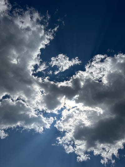The media file is a photograph of the sky, featuring dramatic clouds and blue sky. The clouds are a mix of bright white and dark gray, suggesting a dynamic weather pattern. Sunlight is visible filtering through some of the clouds, creating crepuscular rays that fan out across the sky. These rays are particularly noticeable in the upper left portion of the image and in the right portion. The overall impression is one of a bright and possibly windy day. There are no people or identifiable objects in the frame, and no visible text. The scene is an outdoor sky view, likely taken in Skovorodino, Russia. The presence of both bright and dark clouds indicates it could be mid-afternoon, with the sun still high in the sky. The strong contrast suggests a dramatic sky, possibly before or after a rain shower.