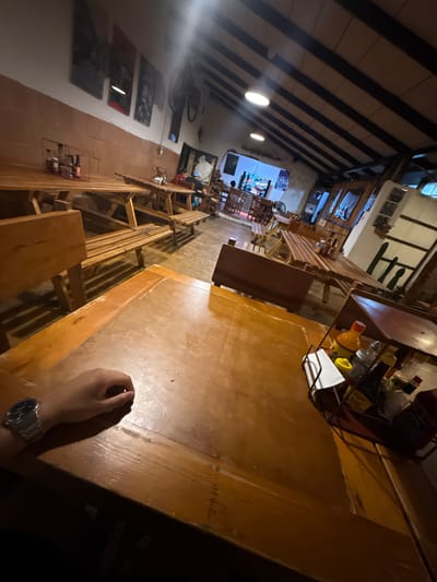 A restaurant interior in Chipilo de Francisco Javier Mina (Chipilo), Mexico. The scene captures the perspective of a person seated at a long wooden table. There are other wooden tables and benches lining the sides, stretching back into the distance. Condiment containers sit on the table. The ceiling features exposed beams. A person's hand and wrist are visible in the foreground wearing a watch.