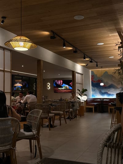 The image depicts the interior of a restaurant located in Tuxtla Gutiérrez, Mexico. The space is characterized by a blend of modern and rustic design elements. The ceiling and walls are adorned with wood paneling, interspersed with track lighting and decorative light fixtures. A large mural, depicting what appears to be a landscape, is visible along one wall.
The dining area features tables and chairs, primarily made of wicker and wood. Several people are seated at the tables, suggesting a populated dining environment. The activity levels suggests the restaurant is open for service, likely in the evening or at night, given the lighting. A television screen displaying food is fixed on the wall.