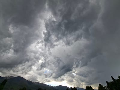 The image captures a dramatic sky over a mountainous landscape in Toguz-Bulak, Kyrgyzstan. The primary subject is the sky, filled with dark, heavy storm clouds that dominate the frame. Streaks of lighter clouds and hints of blue sky peek through in places, suggesting a brewing storm or a break in the weather. The light is diffused and moody, indicative of overcast conditions, likely late afternoon or early evening.

Below the turbulent sky, a range of mountains forms the horizon. The peaks are dark and silhouetted against the lighter sky, with some appearing to have snow on their upper reaches. In the foreground, the tops of dark green trees are visible on the right and left edges of the frame, their silhouettes sharp against the sky. The overall scene evokes a sense of impending weather change, perhaps a thunderstorm. There are no people or explicit objects, making the natural landscape and the dramatic sky the focus. The setting is clearly mountainous, with the lush green of the trees suggesting a valley or lower elevation area. The mood is somewhat somber and powerful, due to the intense cloud formations and the looming mountains. There is no visible text in the image.