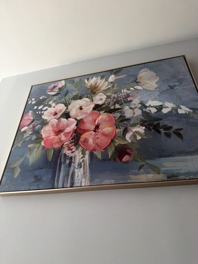 The image shows a framed floral painting hanging on a white wall. The painting depicts a bouquet of flowers in shades of white, pink, and red against a dark blue background. The flowers appear to be painted with thick brushstrokes, giving them a textured appearance. The frame is thin and metallic. The location is not discernible from the image.