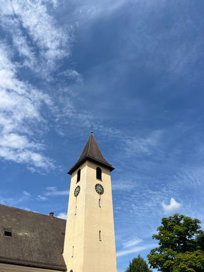 This media file is an upward-facing shot of a church tower and the sky. The tower is light yellow with a dark brown, pointed roof topped with a cross. It has two visible round clocks with green faces and Roman numerals, as well as several rectangular openings. Part of a brown-tiled roof of an adjacent building is visible in the foreground.
The sky is a vibrant blue, dotted with wispy white clouds. A large tree with green foliage is partially visible in the upper right corner. The image appears to have been taken during the day, likely in the late morning or early afternoon, given the bright sunlight and clear sky. The overall mood is peaceful and serene, suggesting a calm, pleasant day. There are no people visible in the image. No text is discernable. The context provided indicates the location is Baden-Baden, Germany.