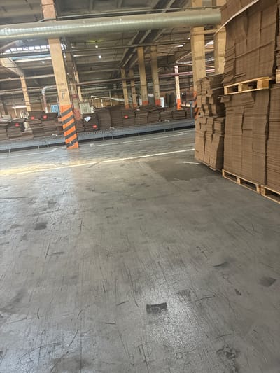 This is a medium-shot, eye-level, indoor photograph of a large industrial warehouse. The floor is concrete and appears worn with scuff marks and stains. In the foreground, the floor takes up the majority of the frame. In the mid-ground, there is a conveyor belt system running horizontally across the image. Stacked cardboard boxes are visible on either side of the conveyor belt, with the stacks on the right being much taller. One stack on the right is elevated on wooden pallets. A support pillar painted with black and orange diagonal stripes stands on the left side of the conveyor belt. Beyond the conveyor belt, the warehouse interior is visible, with more stacked cardboard and industrial infrastructure including pipes, lighting, and support beams. The overall lighting suggests it is daytime. There are no people visible in the image. The location is identified as the Kashira Paper Mill in Kashira, Russia.