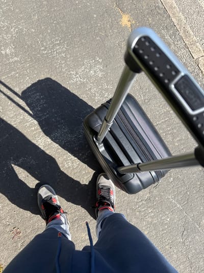 A person stands on a grey paved surface in Welwyn Hatfield, United Kingdom, pulling a black "FLIGHT" branded carry-on suitcase with a silver extendable handle. The person wears grey and black sneakers with red accents and blue pants. The scene is illuminated by bright sunlight, casting distinct shadows of the person and the suitcase.