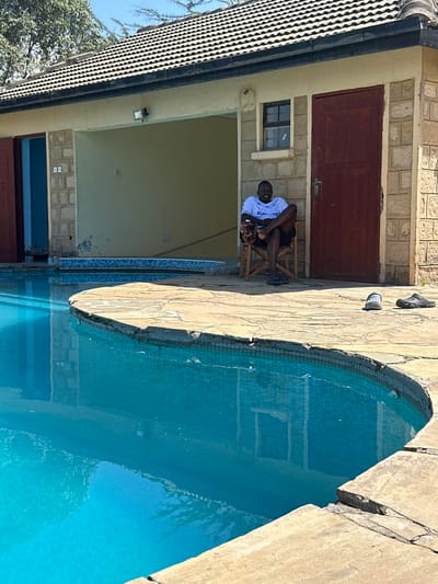 The image depicts an outdoor scene in Olkaria ward, Kenya, featuring a single adult male positioned beside a swimming pool, in front of a single-story building. The male, wearing a white "SUPA-BET" t-shirt, dark shorts, and dark flip-flops, sits on a wooden chair, facing the viewer with crossed ankles and hands clasped.
The building behind him has a dark tiled roof, light-colored stone and plaster walls, and includes a large open archway, a small window, and two dark-red wooden doors, one of which is partially open. An exterior floodlight is affixed above the archway.
A curved swimming pool, filled with clear blue water, occupies the foreground and midground, reflecting portions of the building. The pool is surrounded by an irregular flagstone deck, where two additional pairs of footwear (one light, one dark) are placed to the right of the seated individual. The scene is illuminated by bright sunlight, with sparse vegetation visible in the distant background.