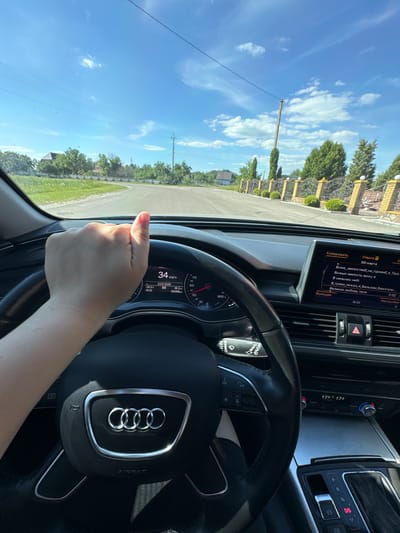This media file shows a first-person perspective from inside a car, likely an Audi, as indicated by the prominent logo on the steering wheel. The driver's hand is visible on the steering wheel, suggesting they are driving. The dashboard displays various information, including the speed (34 km/h), date (June 23, 2026), outdoor temperature (+26.5°C), and time (16:53). The car's infotainment system is also visible, displaying a list of media or song titles in Ukrainian, such as "Боже, милостивий негідний я Твої" and "Великая любовь твоя".
The scene outside the car, visible through the windshield, depicts a bright, sunny day with a clear blue sky dotted with white clouds. Green trees and a fence are visible along the roadside, suggesting a rural or suburban setting. The presence of a utility pole and power lines further indicates an outdoor environment. The overall atmosphere appears to be pleasant and relaxed, consistent with a daytime drive on a summer day. The image is oriented vertically, implying it was taken with a phone held in portrait mode.