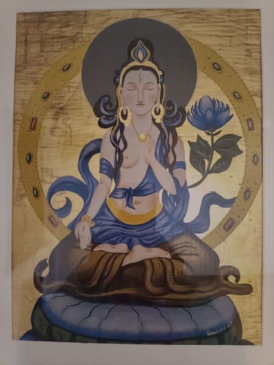 This image depicts a framed painting of a deity, likely a Bodhisattva or similar figure, rendered in a stylized manner. The figure is seated centrally in a cross-legged meditative pose, with eyes closed and a third eye depicted on the forehead. The figure has light skin, long dark hair adorned with an elaborate blue and gold headdress, large golden hoop earrings, a golden necklace, and golden bracelets on both arms. The upper torso is mostly bare, draped with flowing blue fabric, while the lower body is covered by blue and gold garments. The right hand is raised in a gesture, and the left hand rests in the lap.
The figure is seated on a multi-layered blue and green lotus pedestal. Behind the head is a solid dark blue circular halo, and a larger, textured golden circular mandorla with embedded circular and rectangular decorative elements surrounds the entire figure. To the right of the figure, a blue lotus flower with green stem and leaves is depicted. The background of the painting is a textured golden surface. A signature "SURIVALIEV 2018" is visible in the bottom right corner of the painting. The artwork is contained within a simple white frame. There are no discernible features within the image to indicate a specific physical location in the city of Jérica, Spain. The image itself is a digital photograph of this painting.