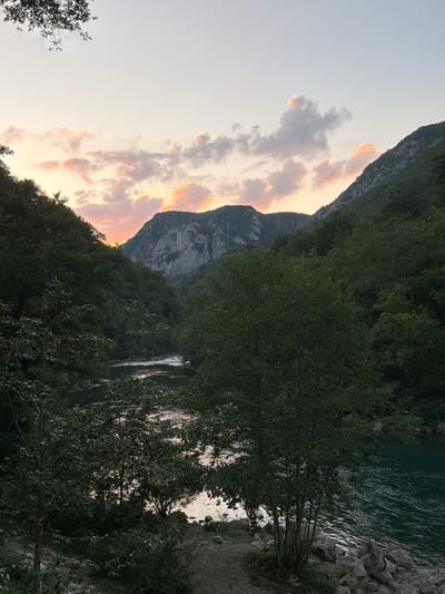The image depicts a serene natural landscape at dusk, likely located in the vicinity of Brijeg, Montenegro. In the foreground, a dense forest lines the banks of a wide river, its surface reflecting the soft hues of the setting sun. The water is a deep emerald green, with ripples indicating a gentle current. Rocky outcrops and a dirt path are visible along the riverbank, partially obscured by low-lying vegetation and the trunks of several trees.

Beyond the river, a dramatic mountainous terrain rises steeply. The slopes are covered in dark green foliage, transitioning to exposed, grey rock faces higher up. The peaks of these mountains are silhouetted against a pale, dusky sky. Scattered clouds in the upper atmosphere are tinged with soft pink and orange, suggesting the last light of sunset. The overall atmosphere is peaceful and tranquil, capturing the essence of a remote, natural environment. There are no people or animals visible in the image.