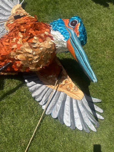 An elaborate, large-scale puppet of a bird is lying on a patch of vibrant green grass. The puppet is intricately crafted with a variety of textures and colors. Its body is covered in overlapping, scale-like elements in shades of burnt orange, gold, and tan, giving it a feathered appearance. The head is adorned with bright blue, textured feathers and has a prominent eye with a dark pupil. The beak is long and pointed, painted in a striking turquoise and orange pattern. One wing is visible, spread out on the grass, displaying large, gray, feather-like segments edged in a light blue. A thin, wooden pole extends from the body of the puppet, resting on the grass. Another dark, cylindrical object is attached to the upper body of the puppet. The overall impression is that of a detailed and colorful artistic creation displayed outdoors. The location is specified as Leominster, United Kingdom.