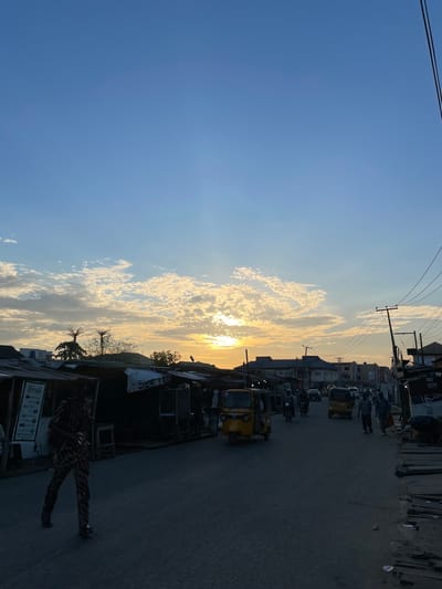 The image shows a street scene in Isheri, Nigeria, at what appears to be dusk or dawn. The sky is visible with white clouds as the sun sits low on the horizon. The road stretches into the distance with buildings and power lines visible on the right. There are vehicles in the street, including small commercial auto rickshaws (tuk-tuks) and potentially cars and motorcycles. There are also people on the street, and one person is particularly visible in the foreground, along with storefronts and signage. The lighting suggests it is either sunrise or sunset, with silhouettes becoming pronounced.