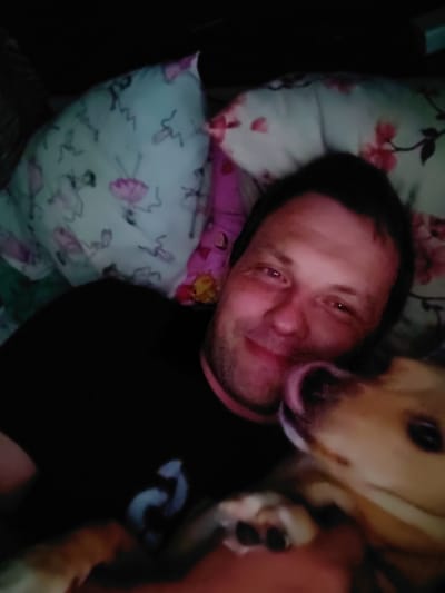 A man and a dog are lying in bed. The man is on his left side, facing the camera. He is wearing a black t-shirt. The dog is on its back, facing the camera with its head near the man's face. The dog is light brown and white. The background consists of pillows with floral patterns. The scene is dimly lit. The location of this image is in Андрэеўшчынскі сельскі Савет, Belarus.
