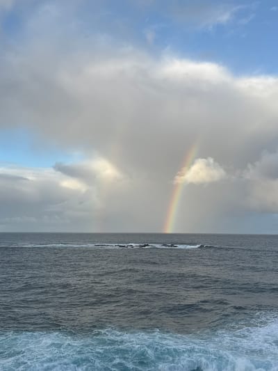 A dramatic sky with a rainbow arc is captured over a dark, choppy ocean. The rainbow, a vibrant spectrum of colors, is partially obscured by soft, gray clouds. Below, the water displays a range of dark blues and grays, with white foam cresting the waves in the foreground. A low, rocky outcrop is visible in the middle distance, breaking the surface of the water. The overall scene conveys a sense of natural power and beauty. The location is identified as Clovelly, Australia, implying an outdoor coastal environment.