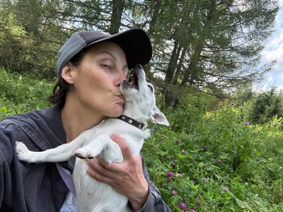 An outdoor selfie captures a woman wearing a black baseball cap with a white checkered brim and a dark gray zip-up hoodie. She is leaning in to kiss a small white dog with black markings around its eyes and ears. The dog is wearing a black collar with silver studs and is being held by the woman. They are positioned in the foreground, with a lush green environment of trees and wildflowers in the background. The location is identified as Lipovaya I, Russia.