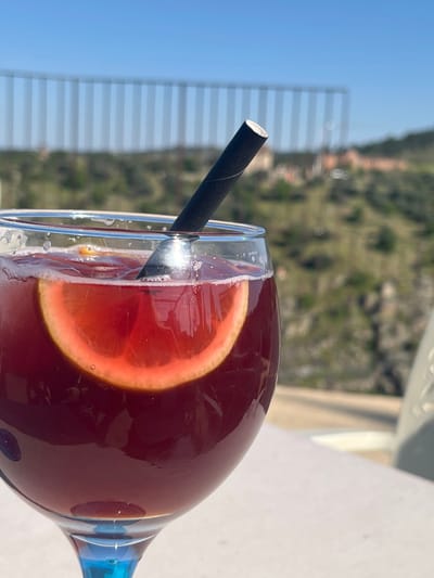 A close-up, outdoor shot depicts a glass containing a dark red beverage, possibly sangria, garnished with a crescent-shaped orange slice and a black paper straw. The drink's surface shows foam and small bubbles. The glass, featuring a blue-tinted stem, rests on a light-colored, flat table surface. A section of a light-colored chair is visible on the right. No individuals are present, and no discernible actions or interactions are depicted. The background, intentionally blurred, reveals a clear blue sky above a prominent, long, light-colored viaduct or bridge structure with numerous vertical supports. Below this structure are green, vegetated hills. In the far, indistinct background, buildings with reddish roofs are faintly visible. This scene is situated in the Toledo, Spain area, specifically from an elevated viewpoint overlooking a valley, likely the Tagus River valley, characteristic of its hilly landscape and modern infrastructure.
