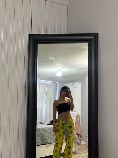 A woman is reflected in a rectangular mirror with a black frame mounted on a white paneled wall. She is wearing a black strapless top and yellow pajama pants adorned with cartoon faces. She holds a phone, presumably taking the picture. The mirror reflects a room with white walls, a white door, curtains, and bedding. A light fixture is visible on the wall behind the woman.