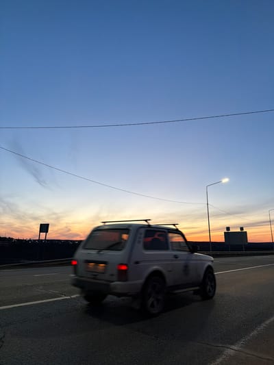 The image depicts a gray or white SUV driving on a road at dusk or dawn. The setting appears to be near the edge of a city, with a row of lights suggesting urban development on the horizon. The sky is a gradient of blue to orange and yellow. There are signs present, and a single lamppost is visible. The location is Kuregovo, Russia.
