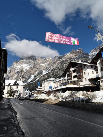 The scene depicts a street view in the city of Serdes, Italy, under clear, sunny conditions with scattered white clouds against a deep blue sky. Snow-capped mountains dominate the background, with jagged peaks and visible rock formations.

A pink banner hangs horizontally across the upper portion of the street, reading "Welcome to San Vito di Cadore Dolomiti Bellunesi" in white text, followed by "Ciao!" and an Italian flag motif.

The main thoroughfare is an asphalt road, appearing wet or damp in sections, with snow banks accumulated along both sides. On the left, a dark multi-story building frames the foreground, followed by a white church with a tall steeple, a visible clock face, and a snow-covered roof. A single evergreen tree stands beside the church. Two passenger vehicles, one dark and one lighter-colored, are visible on the road in the mid-distance near the church.

On the right side of the street, multiple multi-story buildings are present, predominantly white with dark wooden architectural elements such as balconies and rooflines. These buildings also have snow on their roofs and ledges. A blue sign with a white pedestrian crossing symbol is affixed to a pole on the right sidewalk. A black streetlight pole with a white star-shaped decorative fixture is visible on the far right. No people are present in the scene.