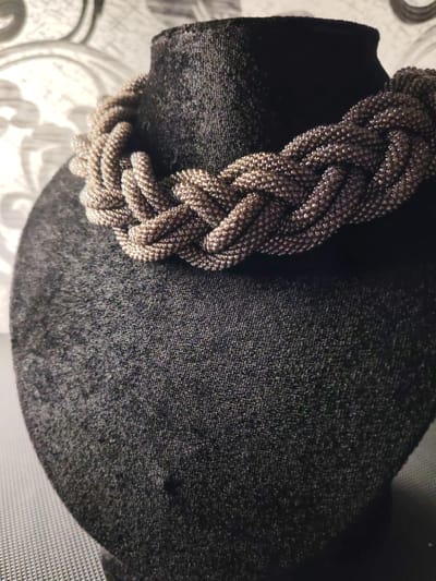 A beaded necklace with a braided design is displayed on a dark fabric bust. The necklace is a metallic, bronze color and appears to be made of tiny beads. The bust is covered in a textured, dark fabric, possibly velvet or velour. Behind the bust, a wallpaper with an abstract, swirling pattern in shades of grey and white is visible. The lighting is soft, creating subtle shadows and highlighting the texture of the necklace and bust. The image appears to be a product shot or a display of jewelry. There are no people visible in the image. The scene does not provide any specific information about the city of Сокол, Russia.
