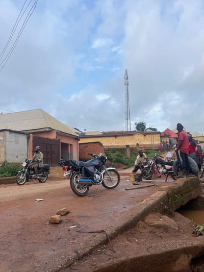 The image depicts a scene in Gembu, Nigeria, characterized by a collection of motorcycles and people gathered on a dirt road. The atmosphere appears to be a typical day, with overcast skies and a generally muted color palette dominated by browns and grays, suggesting a cloudy or possibly rainy day.
Several motorcycles are parked or in use. In the foreground, a person is seated on a black motorcycle, wearing a light-colored shirt and a traditional cap. Further up the road, another motorcycle, this one black with blue accents and a red emblem, is partially visible, with a rider also on it. In the background, a group of people are clustered around motorcycles, some standing and others seated. They are dressed in casual clothing, with one individual in a red shirt and another in a striped garment.
The setting is rural or semi-urban, with buildings visible in the background. One building has a corrugated metal roof and light-colored walls. Another structure further back appears to be made of brick. A prominent feature in the distance is a tall telecommunications tower, indicating a degree of modern infrastructure within the locale. The road itself is unpaved, with areas of mud and scattered debris, common in many Nigerian towns and villages. The overall impression is one of everyday life, with motorcycles serving as a primary mode of transportation and a social gathering point. No discernible emotions are evident from the individuals depicted, as the focus is on the static scene. There is no visible text that is clear enough to be read.