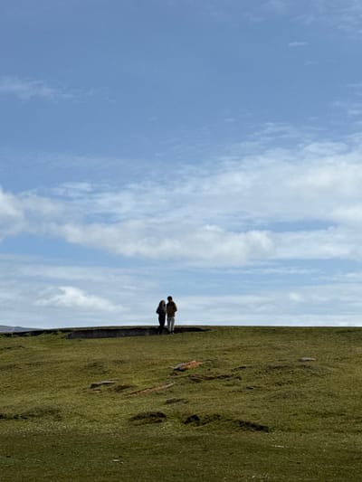 An outdoor wide shot depicts a vast, open landscape at Bluff Cove, Falkland Islands (Islas Malvinas), under a partly cloudy blue sky. The upper two-thirds of the frame is dominated by a clear blue sky interspersed with scattered white cumulus clouds, more concentrated towards the left and lower horizon. The lower third features a uniformly green, short-cropped grassy terrain with subtle undulations and darker patches. Several elongated, reddish-brown objects, possibly rocks or debris, are sparsely distributed on the grass in the mid-left. Two individuals stand near the center of the image, silhouetted against the sky on the horizon line. The person on the left wears dark clothing, and the person on the right wears lighter, possibly tan, attire. They are standing in close proximity, appearing to face towards the right side of the frame. A dark, low-lying linear feature is visible directly beneath them, at the edge of the elevated ground they occupy. The scene suggests a remote, natural environment.