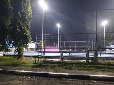 This media file captures an outdoor sports court at night, illuminated by bright floodlights. The scene is a sports facility, likely a tennis or basketball court, given the blue surface and visible boundary lines. A chain-link fence encloses the court, with metal bleachers positioned outside the playing area.
Several figures are visible on the court, suggesting an ongoing activity. One person is seen running, while others are standing or walking, hinting at a practice session or a casual game. The lighting creates strong highlights and shadows, typical of nighttime illumination. A large tree with dense foliage stands to the left of the frame, partially obscuring the view of the court. Beyond the court, a building with blue and pink accents is visible, with some text on it that is not fully legible but appears to be in Indonesian, possibly indicating the location or facility name.
The time of day is clearly night, with a dark sky. There are no visible indicators of the weather, though the clear sky suggests it might be fair. The overall atmosphere is one of activity and sporting engagement, even in the late hours, facilitated by the powerful artificial lighting. The location context provided is Palohgadeng, Indonesia.