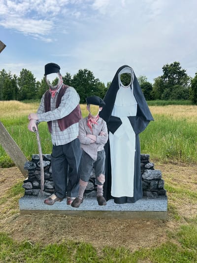 This media file captures a tableau of three mannequins depicting figures dressed in historical attire. The central figure is a nun in a traditional black and white habit, standing behind two other figures. In front of the nun, a man dressed as a farmer, wearing a plaid shirt, waistcoat, and cap, leans forward holding a pitchfork. To his right, a boy in striped trousers and a simple shirt, also wearing a bandana, is positioned as if being carried or supported by the farmer. The mannequins are arranged on a stone base, set against a backdrop of green fields, dry grass, and trees under a cloudy sky. The scene appears to be outdoors, possibly in a rural setting in Zichem, Belgium, given the location context. The overall impression is one of a historical depiction, perhaps representing a scene of charity, care, or hardship from the past. The lighting suggests it is daytime, but the specific time is not discernible. There is no visible text in the image.