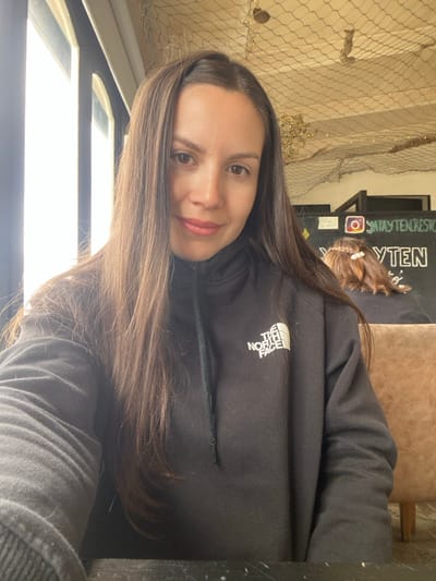 The image captures a female individual seated indoors at what appears to be a restaurant in Puerto Almanza, Argentina. The woman, estimated to be in her late 20s to early 30s, possesses long, straight dark brown hair, light brown eyes, and pink-toned lips, and is looking directly at the camera with a slight smile. She is wearing a black hooded sweatshirt with a zipper and drawstring, featuring the white "THE NORTH FACE" logo on the left chest. She is positioned behind a dark, reflective surface, likely a table.
To her left, a large window with vertical dark frames allows significant natural light to enter. The background to her right reveals a commercial interior. A dark wall or signboard displays the text "@SANTAYTENRESTO" next to an Instagram logo, identifying the establishment as "Santa Y Ten". Below this, "YTEN" is partially visible in larger white text. Another individual is seated further back, visible from behind, with brown hair styled in a braid adorned with small white beads or clips. A light brown, upholstered booth or chair is visible behind this second person. The ceiling is covered with a light-colored decorative fishing net, consistent with a nautical or rustic theme, from which several small, indistinct objects are suspended. The specific identification of "Santa Y Ten" confirms the location within Puerto Almanza, Argentina.