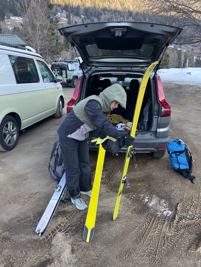 The image depicts an outdoor scene in Vallorcine, France. A single individual, dressed in an olive-green hooded jacket with black sleeves and grey mid-section, black pants, black gloves, and light-colored sneakers, is positioned facing the open rear liftgate of a dark grey SUV. The person is bent over, engaged in handling a pair of bright yellow skis. One ski is held upright, leaning against the vehicle, while its counterpart lies on the unpaved, dirty ground which displays numerous tire tracks and patches of melting snow. A black backpack rests near the individual's feet, and a blue backpack is situated further to the right on the ground. The open trunk of the SUV reveals a coiled rope and other gear. To the left, a cream and white Volkswagen van with a roof rack is parked, and a green Land Rover Defender with a roof rack is visible further in the background. The environment consists of forested mountain slopes with intermittent snow patches under natural lighting conditions.