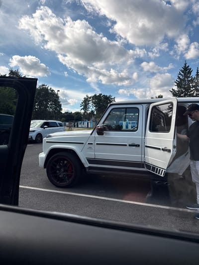 In Zhukovka, Russia, under a partly cloudy sky, a white Mercedes-Benz G-Class SUV is parked in a lot. The driver's side front door and rear door are open, with a man wearing a black t-shirt, light-colored pants, and a baseball cap exiting the vehicle from the rear passenger side. Another white SUV is parked behind it. The scene appears to be a parking lot with trees and some buildings in the background. The perspective is from inside another vehicle looking out through its open door.