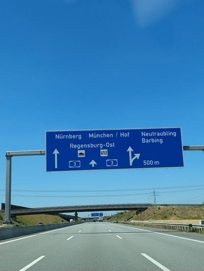 An eye-level, medium outdoor shot captures a blue highway sign against a clear blue sky. The sign provides directions to Nürnberg, München/Hof, and Regensburg-Ost, with an indication of the GVZ (Güterverkehrszentrum, or freight transport center) and a route number 3. Additionally, it directs towards Neutraubling and Barbing, with a distance of 500 meters, and shows an arrow indicating a right turn. Below the sign, a multi-lane highway stretches into the distance, bordered by a concrete barrier and grassy slopes. An overpass is visible in the middle ground, with power lines and a distant transmission tower in the background. The scene appears to be on the outskirts of Barbing, Germany, during daylight hours.